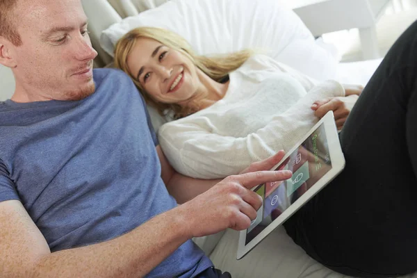 快乐微笑的年轻夫妇躺在床上 在平板电脑上使用智能家居应用程序 — 图库照片