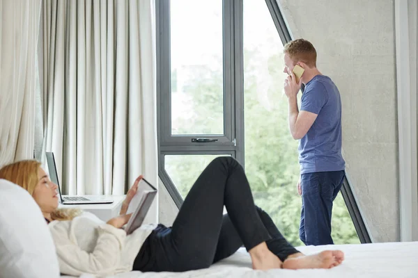 Young man looking through window and talking on phone when his girlfriend lying on bed and reading book