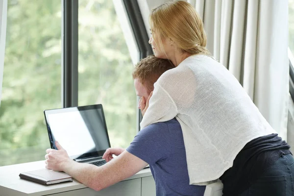 漂亮的年轻女子拥抱她的男朋友与爱 当他检查电子邮件在笔记本电脑 — 图库照片