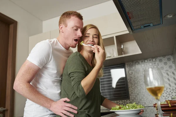 夕食に野菜サラダを作るときに彼氏に食べさせる陽気な若い混合人種の女性 — ストック写真