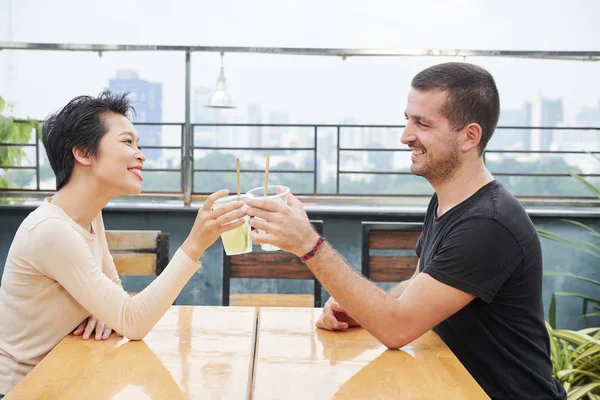 年轻的幸福夫妇坐在对面的桌子边喝着水果鸡尾酒 在户外咖啡馆里微笑 — 图库照片