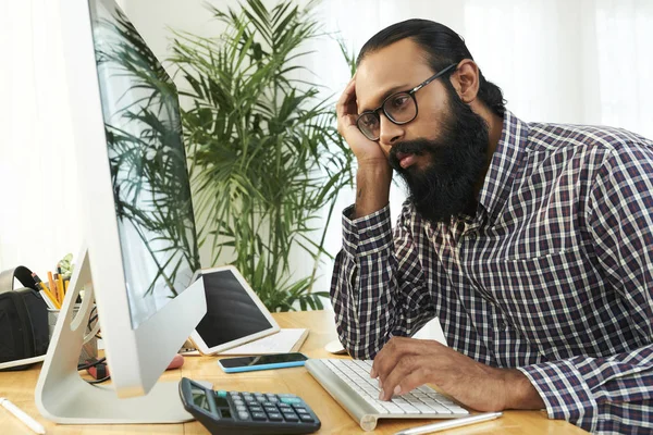 コンピュータのキーボードに入力し 疲れているように見えるコンピュータモニターの前に彼の職場に座って眼鏡でアフリカのひげを生やしたビジネスマン — ストック写真