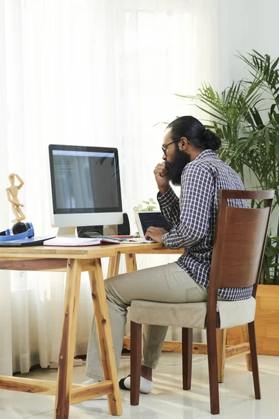 Serious bearded freelancer sitting at the table and examining new software on computer monitor at home