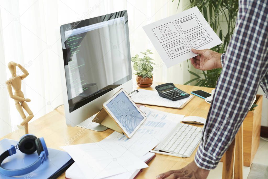 Close-up of programmer holding document with charts and examining it while standing in front of computer monitor with software and digital tablet with business strategy at office
