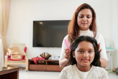 Anne saçlarını tararken ve evde oturma odasında saç modeli yaparken genç kız otururken kameraya bakarken