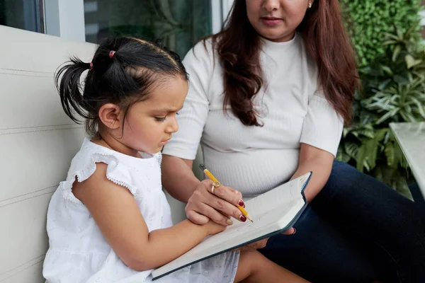 年轻的老师坐在一个小女孩的身上 教她如何在笔记本上用铅笔写字 — 图库照片