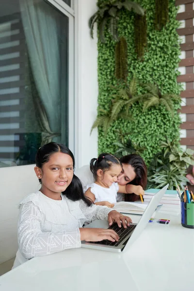 坐在桌旁的女孩在笔记本电脑上打字 在相机前微笑 母亲教妹妹在背景中写作 — 图库照片