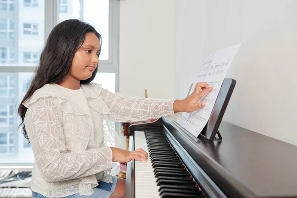 Indian young girl sitting at piano and examining music sheet she learning to play on piano