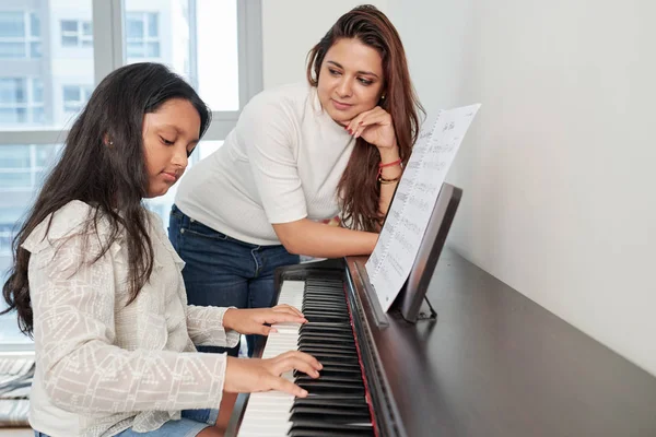 Young woman standing near the piano and looking at her daughter how she playing the piano during musical lesson