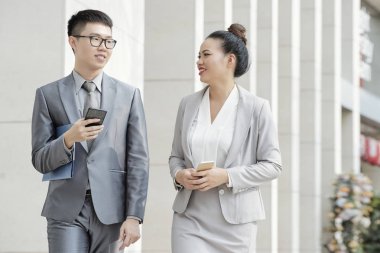 Ellerinde cep telefonu tutan iki iş arkadaşı şehirde yürürken işlerini tartışıyorlar.