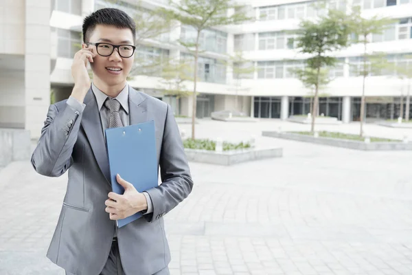 眼鏡をかけたアジアの若いビジネスマンは 青いフォルダを保持し 屋外の街で携帯電話で話すフォーマルな服を着て — ストック写真