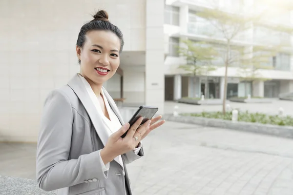 屋外に立ちながら携帯電話を使い カメラに微笑むアジアの若いビジネスウーマンの肖像 — ストック写真