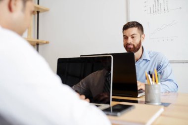 Sakallı genç işadamı işyerinde oturan ve onun önünde meslektaşı ile dizüstü bilgisayarda yazma çok ofiste dizüstü bilgisayar üzerinde çalışan