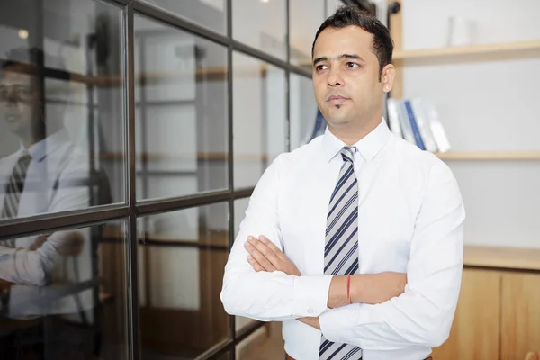 Mature Indian businessman standing in white shirt with his arms crossed at modern office and looking thoughtful