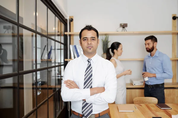 腕を組んで立っているインドのビジネスマンの肖像画が交差し オフィスでバックグラウンドで働くビジネスマンとカメラを見て — ストック写真