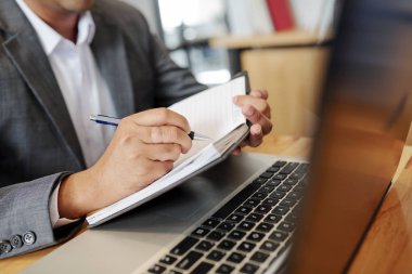 İşinde dizüstü bilgisayar kullanarak masada oturan ve not defterinde kalemle notlar alan işadamının yakın çekim