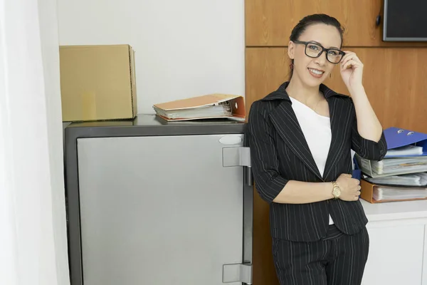 オフィスで働きながら 金庫の近くに立ち カメラに微笑むメガネ姿の女性幹部の肖像 — ストック写真