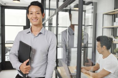 Cam ofis duvarına yaslanmış ve kameraya bakan deri klasörlü pozitif genç finans yöneticisi