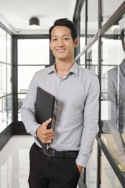Ofis koridorunda duran ve kameraya bakan deri belgeler klasörü ile emin genç Asyalı girişimci