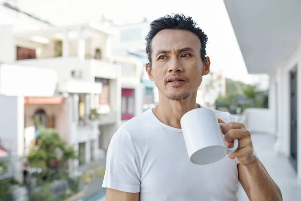 朝バルコニーに立つと黒いコーヒーの大きなマグカップを飲むアジア人男性の肖像 — ストック写真