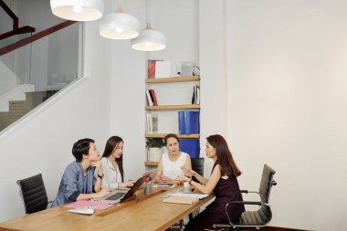 Bazı belgeler ve dizüstü bilgisayarlarla masada oturan ve modern ofiste toplantı sırasında takım halinde tartışan bir grup genç iş kadını