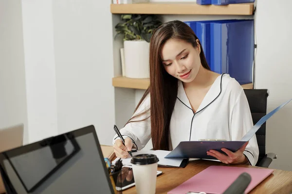 亚洲年轻女商人坐在桌旁 打开文件夹 在办公室工作期间在纸上写点东西 — 图库照片