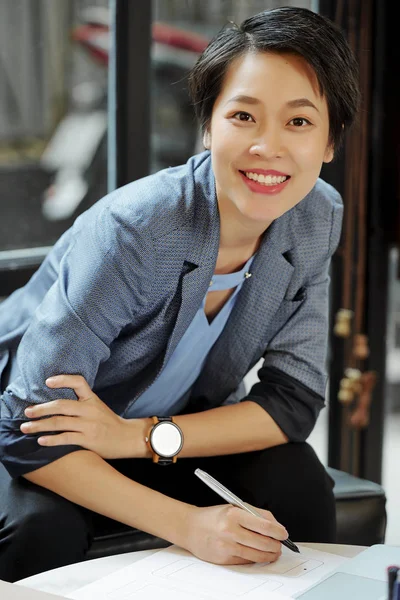 Portrait of Asian manager smiling at camera while sitting at the table and taking notes on paper at office