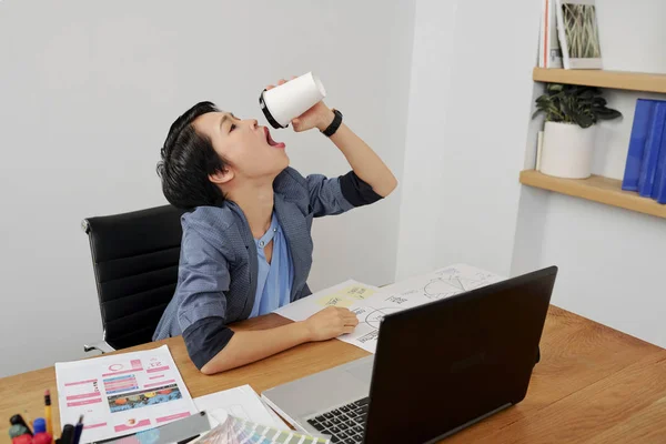 亚洲年轻办公室工作人员坐在她的工作场所与文件和笔记本电脑 喝咖啡从一次性杯到底部 — 图库照片