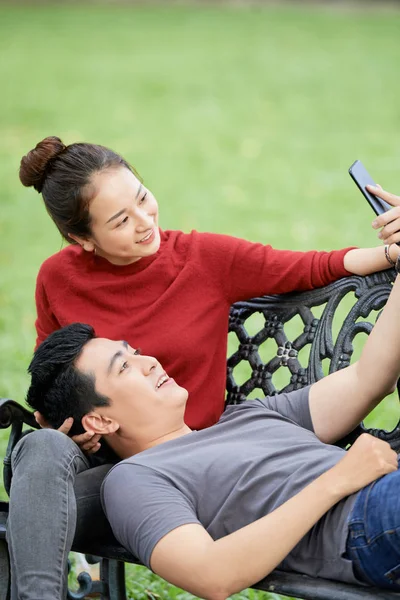 年轻的亚洲男子躺在长凳上 他的头在女朋友的腿上 并采取自拍 — 图库照片