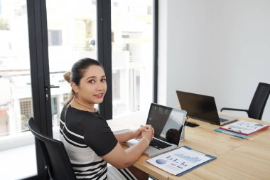 Modern ofiste dizüstü bilgisayar ve finansal grafiklerle işyerinde çalışırken kameraya gülümseyen genç Asyalı iş kadınının portresi