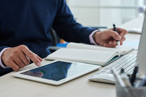 Anonymous man browsing modern tablet and writing in notepad while sitting at desk in office