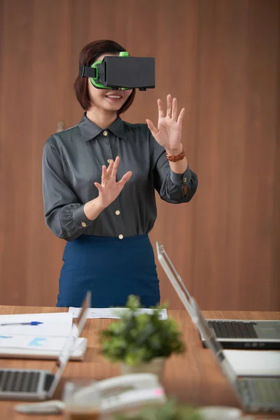 Young businesswoman in virtual reality goggles standing and gesturing, she has virtual business meeting at office
