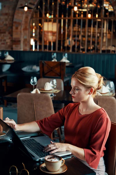 Serious elegant business lady sitting at restaurant table and working on laptop