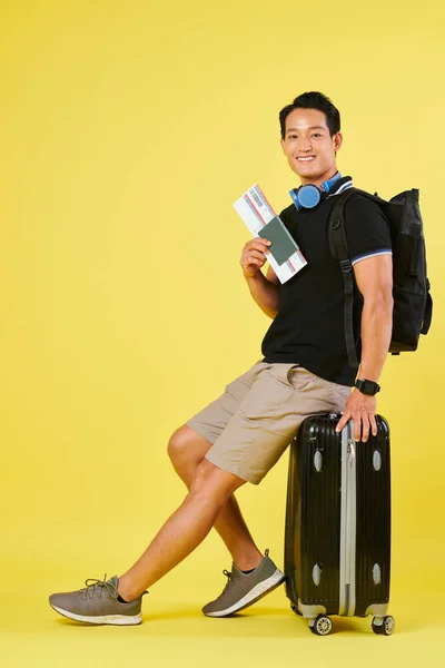 バックパックとチケットをスーツケースに座って搭乗を待っている陽気なアジアの若者 — ストック写真