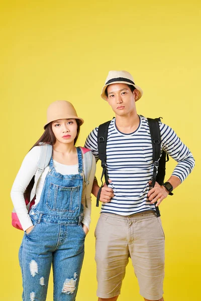黄色の背景に立ってカメラを見てバックパックを持つ観光客の悲しいか退屈な若いアジアのカップルの肖像画 — ストック写真