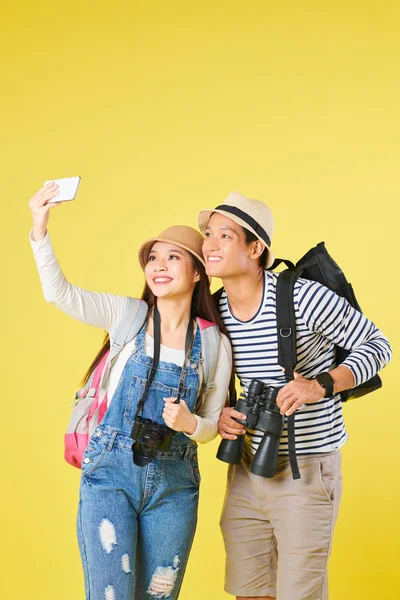 Cheerful young Vietnamese tourists with digital camera and binoculars posing for selfie on smartphone, isolated on yellow