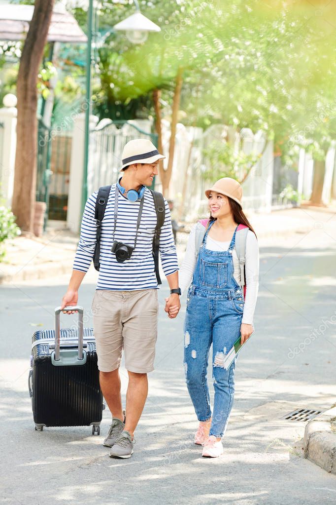Happy young Vietnamese couple holding hands and looking at each other when walking along the road with big suitcase