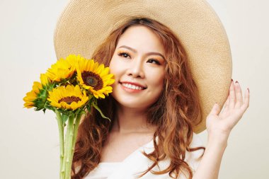 Beyaz arka plan karşı ayçiçeği ile poz verirken gülümseyen şapka giyen kıvırcık kırmızı saçlı Asya moda modeli