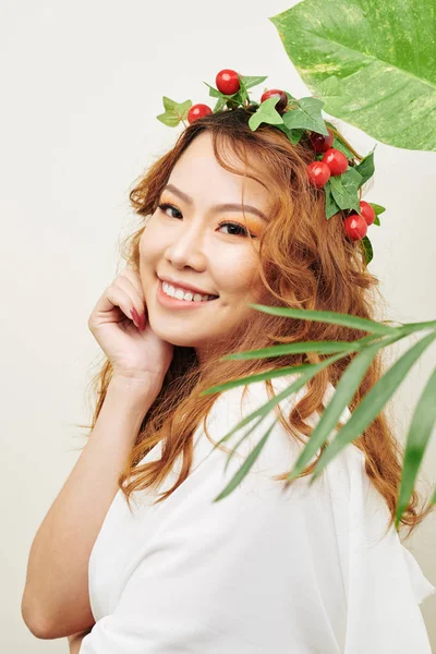 長い赤い巻き毛を持つアジアの若い女性の肖像画とベリーの花輪と彼女の頭の上の葉がカメラで微笑む — ストック写真