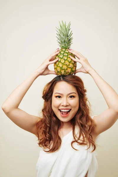 兴奋的亚洲女孩与红色长卷发在她的头上拿着菠萝和微笑在相机孤立的白色背景的肖像 — 图库照片