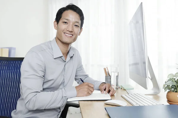 Young smiling Vietnamese entrepreneur writing plans and ideas in his journal when sitting at office table