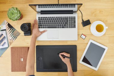 Tablet pc ve dijital fotoğraf makinesi arasında ahşap masada oturan ve ofiste dizüstü bilgisayar önünde grafik tablet kullanarak tasarımcı yakın çekim