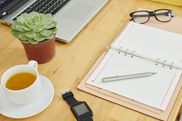 オフィスの木製のテーブルの上にお茶とラップトップコンピュータの白紙のカップと開いたメモ帳のクローズアップ — ストック写真