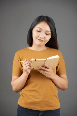 Asyalı genç kadın ayakta ve gri arka plan üzerinde izole onu not defterinde kalem ile planlarını yazma