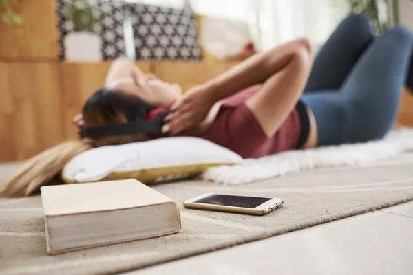 年轻女子躺在地板上 用耳机听音乐 她的智能手机和厚书在前景 — 图库照片