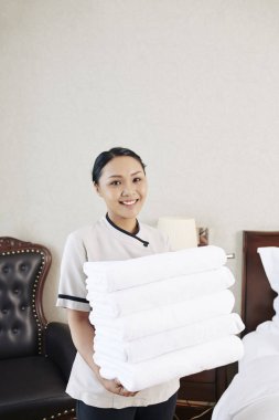 Üniformalı Asyalı genç hizmetçinin otel odasında dururken havlu destesi tutan ve kameraya gülümseyen portresi