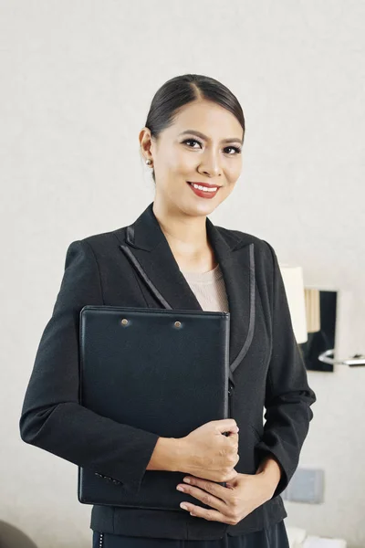 Portrait of Asian young businesswoman in formal black suit holding folder with documents and smiling at camera