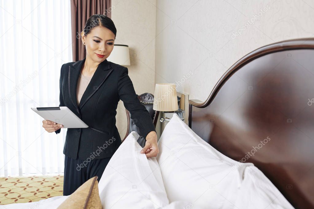 Asian young businesswoman in black suit holding digital tablet and checking the quality of pillows on the bed in the hotel room