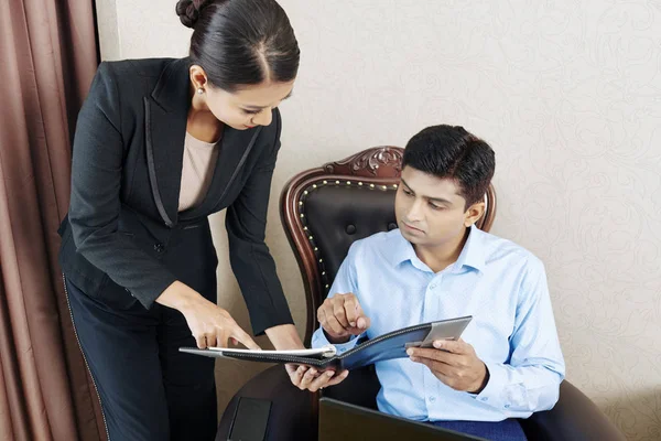 Asian businesswoman in black suit holding folder with business contract and showing it to the young businessman while they working in the hotel room