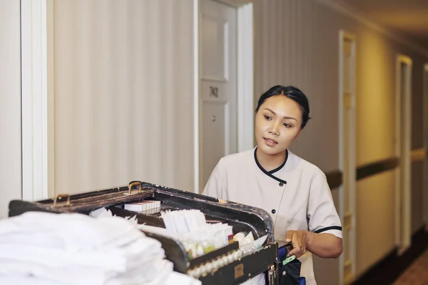 Asian maid in white uniform pushing the cart with different hotel stuff along the corridor of the hotel